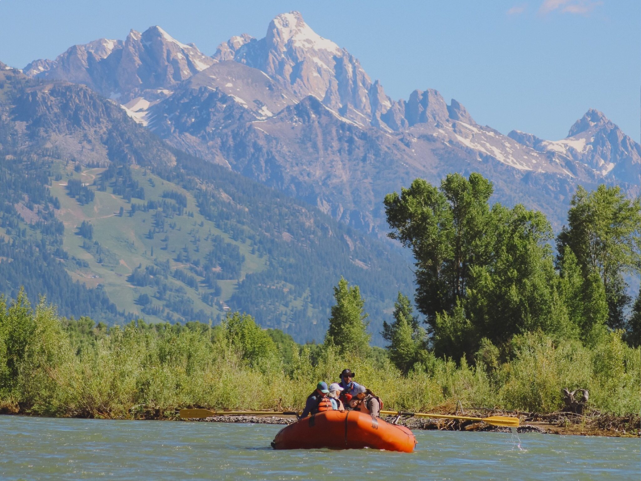 An orange raft carrying a small group floats on a calm river with the jagged, snow-dusted peaks of the Grand Teton mountain range towering in the background. Lush green trees and riverbanks line the water under a clear blue sky.