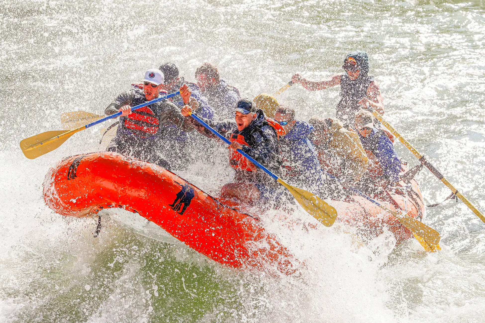 A side profile photo of an orange Lewis and Clark raft splashing through rapids with smiling customers.